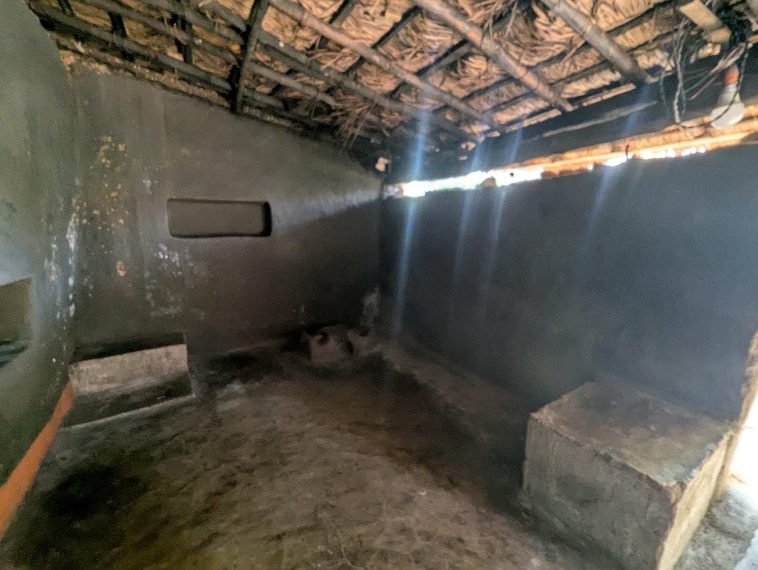 The interior of a regional house: dark earthen walls and floor, a built-in platform, and shafts of light falling from under a timber roof.