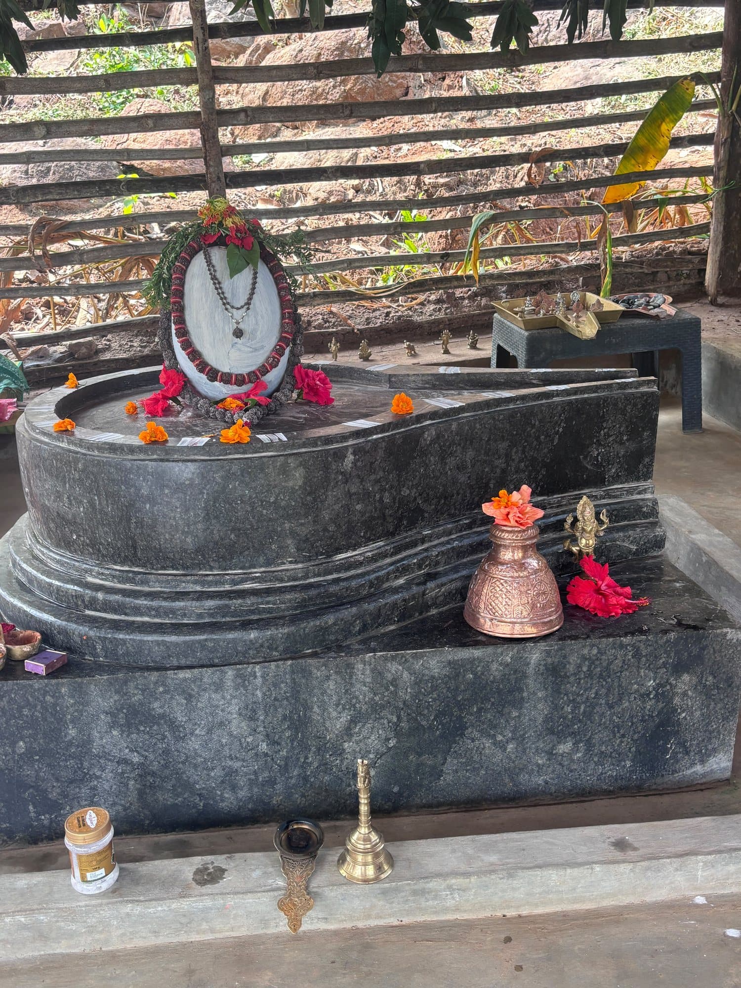 The sacred stone shrine at Shaktigiri: a dark stone pitha with a white stone at its head, garlanded with rudraksha and marigolds, with a copper kalasha, brass bell and lamp in front.
