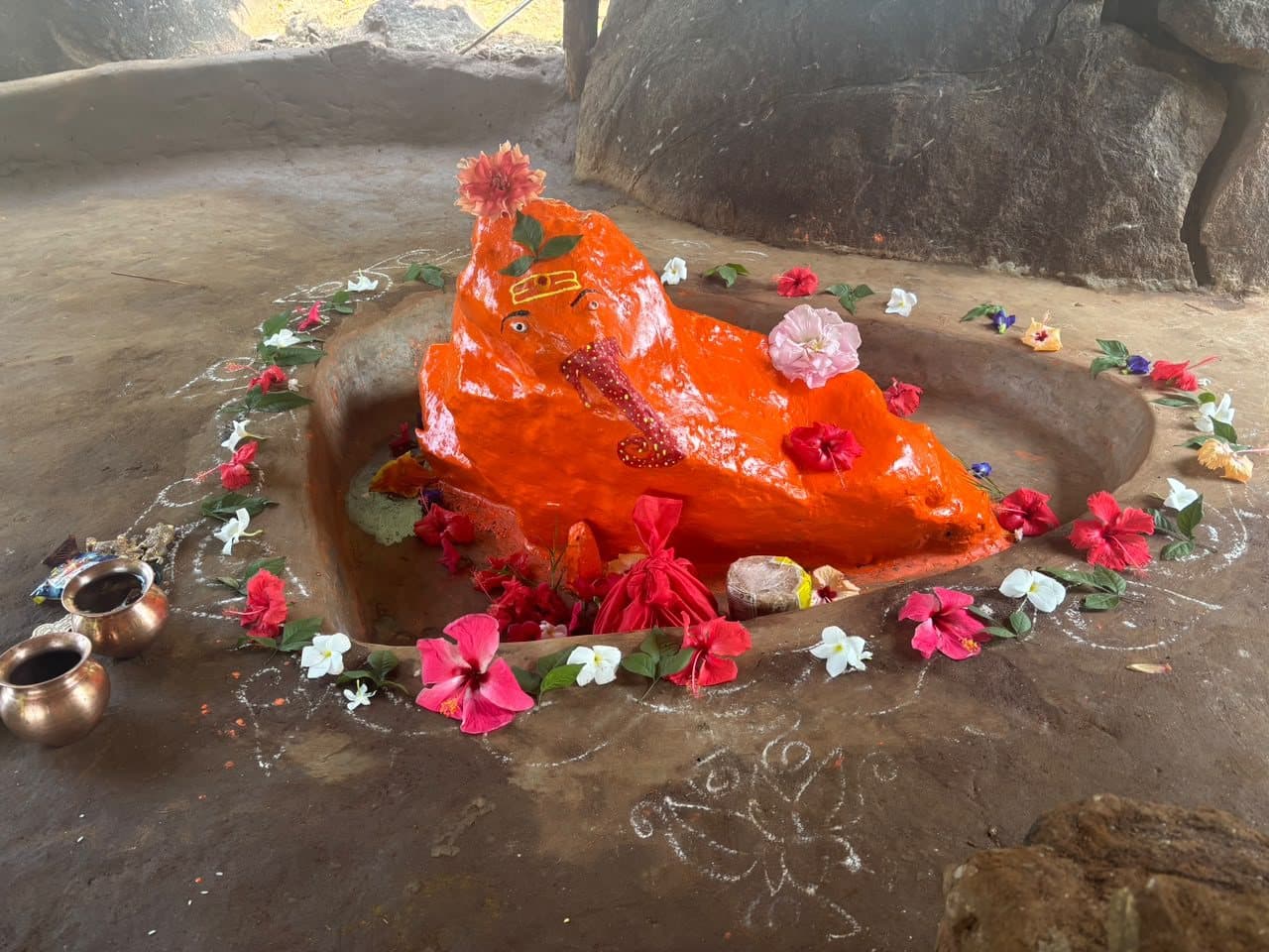 Yoga Ganapati: a natural rock form covered in orange sindoor, resting in a shallow stone basin ringed with hibiscus, frangipani and rangoli, beside large boulders.